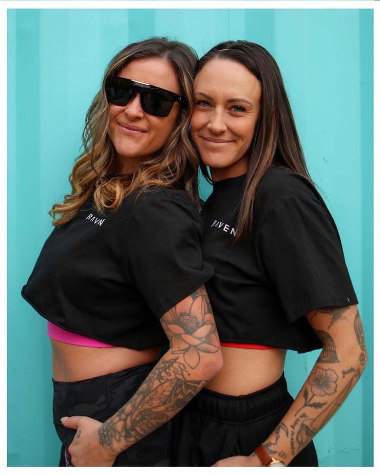 Two models wearing crop tops in black, standing against a blue background, with one model holding the other&